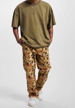 Person iført en olivengrøn oversized t-shirt, orange og brun camouflage cargo bukser, hvide sneakers og et sølvfarvet kædehalssmykke.