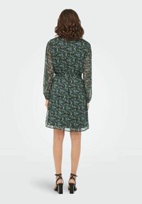 Donna con capelli ondulati lunghezza spalla, che indossa un abito lungo a maniche lunghe con motivo floreale scuro e sandali neri con tacco alto, in piedi di spalle.