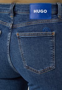 Mørkeblå denimjeans med bakutsikt, med en lærmerke merket "HUGO" og oransje søm på lommene.