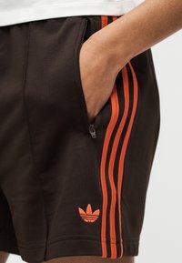 Χέρι στην τσέπη μαύρων σορτς Adidas με πορτοκαλί ρίγες και λογότυπο, φορεμένα με λευκό πουκάμισο.