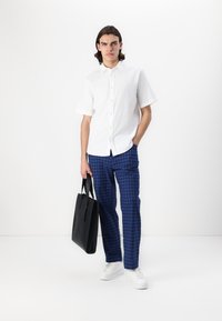 Camicia bianca a maniche corte con bottoni abbinata a pantaloni a scacchi blu. Il modello tiene una borsa a tracolla nera e indossa sneakers bianche.