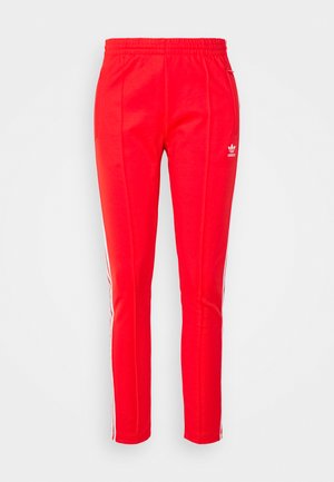 Pantalones deportivos rojos con cintura elástica, corte ajustado y rayas laterales blancas con un sutil logo en el muslo izquierdo. Textura de tela suave.