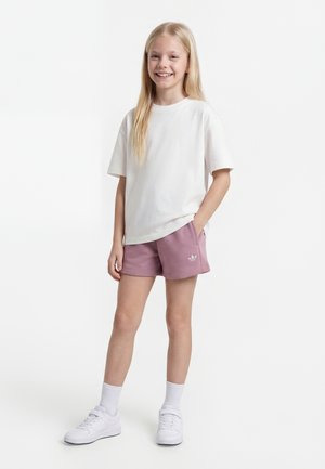 Une jeune fille souriante se tient debout, une main dans la poche, portant un t-shirt blanc, un short mauve avec un logo, des chaussettes blanches et des baskets blanches sur un fond uni.