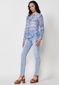 Blusa blu trasparente con motivi geometrici, chiusura a bottoni, maniche lunghe, abbinata a jeans skinny blu chiaro e sandali con tacco in legno marroni.
