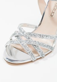 Sandale à talon haut argentée avec des brides croisées pailletées, un bout métallique brillant et une semelle intérieure beige lisse.