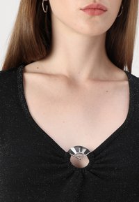 Svart topp med V-halsdesign, med en metallisk sløyfe i midten. Glitrende tekstur, glatt stoff og samlede detaljer ved halslinjen.