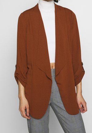 Femme portant un blazer ouvert couleur rouille sur un crop top à col roulé blanc et un pantalon taille haute à carreaux gris.