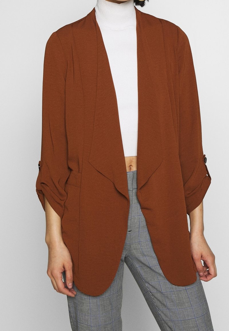Femme portant un blazer ouvert couleur rouille sur un crop top à col roulé blanc et un pantalon taille haute à carreaux gris.