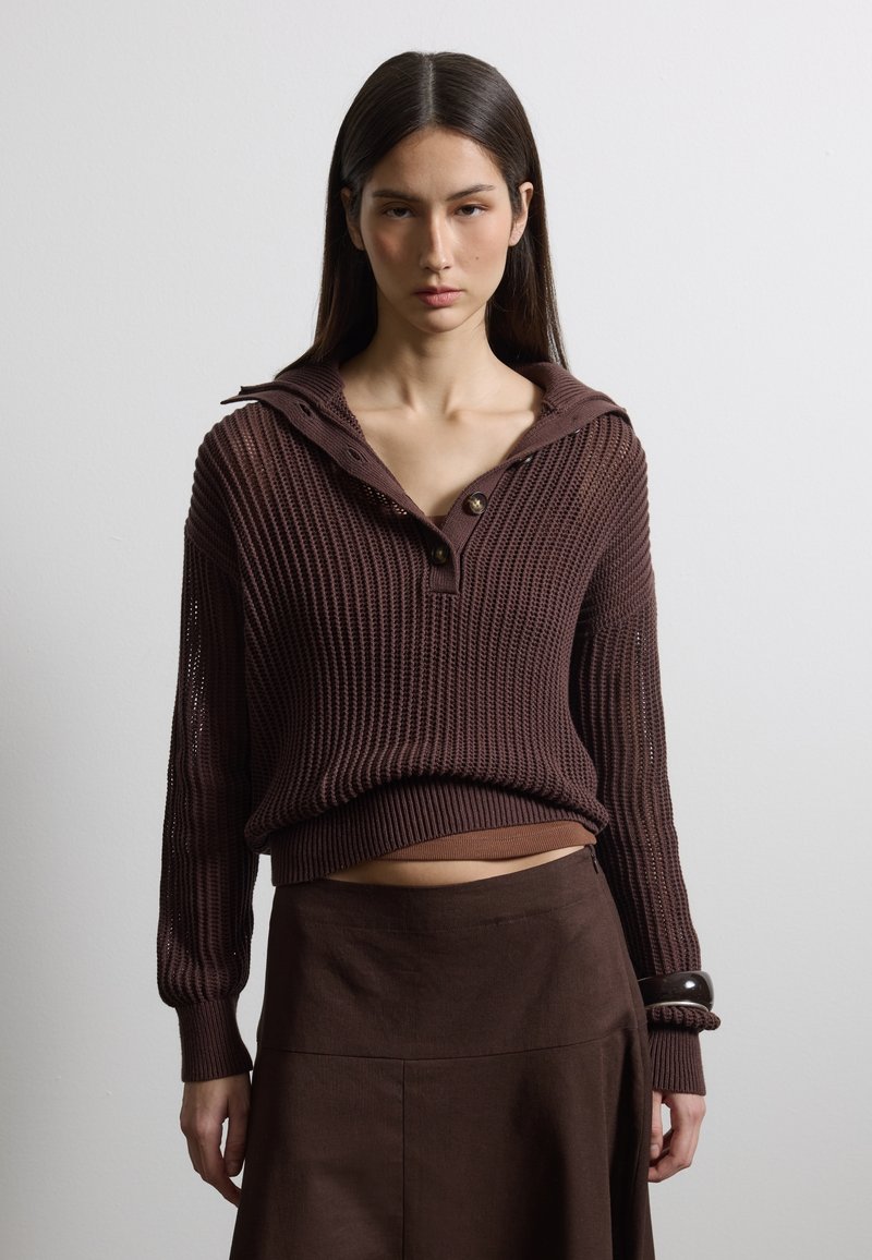 Junge Frau mit langen dunklen Haaren, die einen braunen, gestrickten Pullover mit Knöpfen und einen braunen Rock trägt, vor einem schlichten hellen Hintergrund stehend.