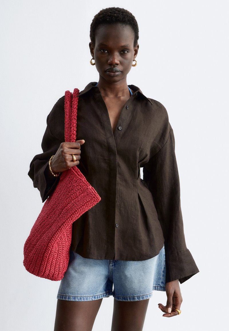 Femme portant une chemise marron foncé boutonnée et un short en jean, tenant un grand sac fourre-tout rouge tricoté sur son épaule.