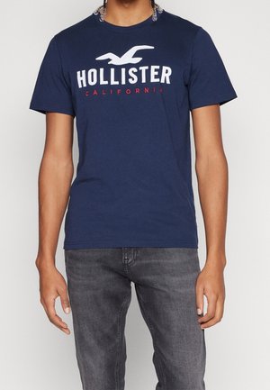 Granatowa bawełniana koszulka z krótkim rękawem z logo "HOLLISTER" w białym i czerwonym kolorze, z okrągłym dekoltem. Gładka faktura, luźny krój.
