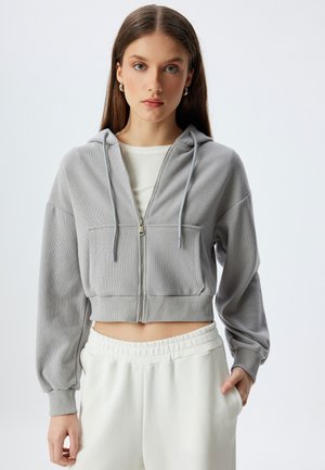 Graue, taillierte Zip-Hoodie aus strukturiertem Stoff, mit einer Fronttasche, Kordelzügen und gerippten Bündchen. Kombiniert mit weißen Jogginghosen.