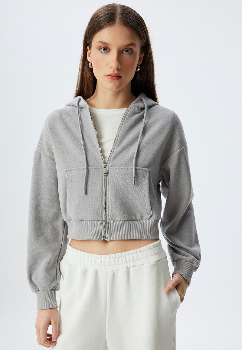 Sudadera gris con cremallera y corte cropped, hecha de tela texturizada, que cuenta con un bolsillo frontal, cordones y puños acanalados. Combinada con joggers blancos.