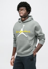 Γκρι φούτερ με κουκούλα και τσέπη καγκουρό, με κέντημα "Air Jordan" σε κίτρινο και λευκό λογότυπο jumpman. Συνοδεύεται από σκούρο παντελόνι με μπαλώματα.