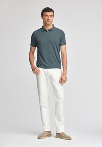 Polohemd in Dunkelgrün, kurzärmelig, mit geripptem Kragen, kombiniert mit weißen Hosen und hellgrauen Espadrilles, stehend vor einem hellen Hintergrund.