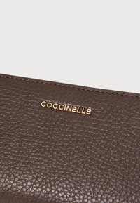 Portofel din piele texturată maro, cu un logo auriu "Coccinelle" afișat proeminent pe față, prezintă detalii de cusătură subtile.