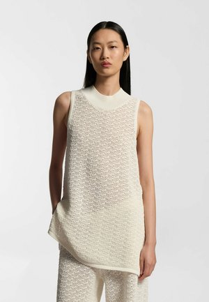 Femme aux longs cheveux noirs portant un haut en maille sans manches texturé blanc cassé et un pantalon assorti, se tenant devant un fond clair uni.