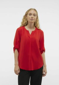 Blusa rossa fluente con scollatura a V e maniche 3/4, caratterizzata da un tessuto testurizzato e un orlo curvo, abbinata a pantaloni neri.