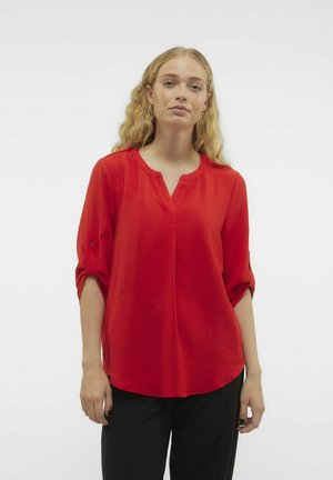 Blouse rouge fluide avec un décolleté en V et des manches 3/4, offrant un tissu texturé et un ourlet incurvé, associée à un pantalon noir.