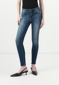 Skinny jeans in dark blue denim, featuring a high waistband, five-pocket design, and raw hem. Paired with black slingback heels.