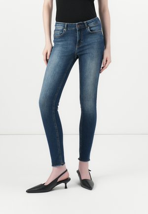 Skinny jeans in dark blue denim, featuring a high waistband, five-pocket design, and raw hem. Paired with black slingback heels.
