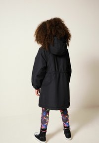 Sort hættetrøje med indsnøret talje, lange ærmer og elastiske manchetter, parret med farverige, mønstrede leggings og sorte sko.