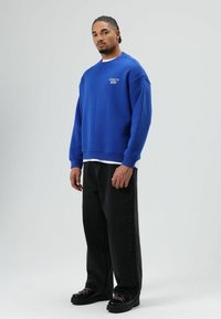 Sweat-shirt bleu avec poignets et ourlet côtelés, porté sur un t-shirt blanc. Jean noir à jambes larges et chaussures noires avec lacets roses. Marque minimale visible.