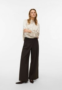 Licht beige blouse met geplooide manchetten, gecombineerd met zwarte wijde broek met hoge taille en donkere hakken. Minimalistisch ontwerp, gladde textuur.