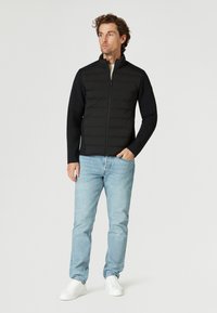 Svart puffjacka med hög krage och dragkedja fram, kombinerad med svarta långärmade ärmar. Buren tillsammans med ljusblå jeans och vita sneakers.
