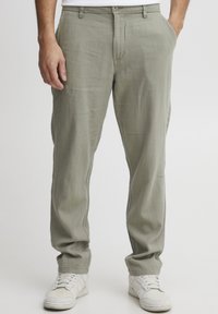 Light green linen trousers feature a straight fit, side pockets, and a button closure. Paired with white athletic shoes.
