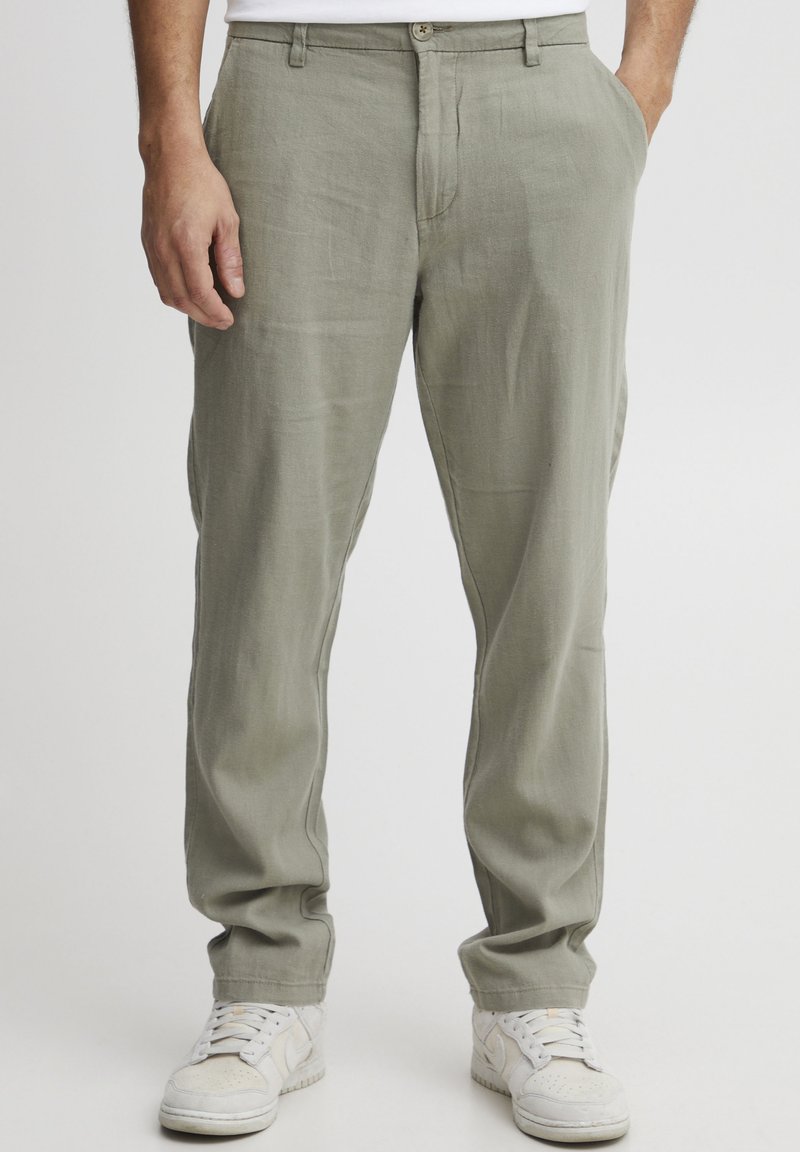 Light green linen trousers feature a straight fit, side pockets, and a button closure. Paired with white athletic shoes.