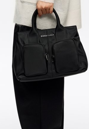 Bolso tote de nailon negro con dos bolsillos frontales con cremallera, logo en relieve en la parte superior, textura suave y asas redondeadas.