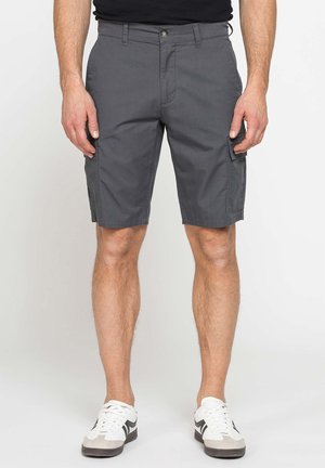 Uomo che indossa pantaloncini cargo grigio scuro e sneakers bianche, in piedi davanti a uno sfondo chiaro uniforme.