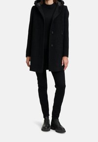 Cappotto in misto lana nero con cappuccio, superficie texturizzata, chiusura con bottoni e tasche laterali, abbinato a pantaloni neri aderenti e stivaletti.