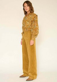 Blouse à manches longues avec motif paisley en couleur moutarde, épaules froncées, associée à un pantalon en velours côtelé taille haute à jambes larges dans une teinte assortie.