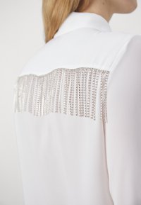 Blusa bianca con colletto e una fila verticale di decorazioni in frange di cristallo sulla parte superiore della schiena, che mette in mostra una texture di tessuto liscia.