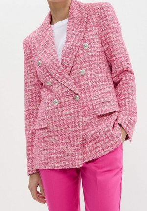 Kobieta ubrana w różowy, dwurzędowy, teksturowany żakiet z srebrnymi guzikami, nałożony na biały top oraz jasnoróżowe spodnie, z ręką w kieszeni.