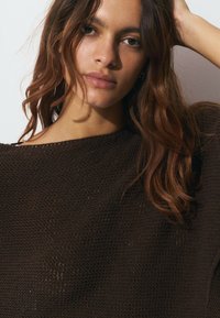 Pull en tricot marron avec une coupe ample et surdimensionnée. Tissu texturé avec des tissages ouverts et un large col bateau. Le modèle pose avec des cheveux en désordre.