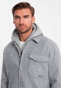 Veste en mélange de laine grise avec un devant boutonné, une poche poitrine et un sweat à capuche gris dépassant en dessous. Texture lisse, design décontracté.