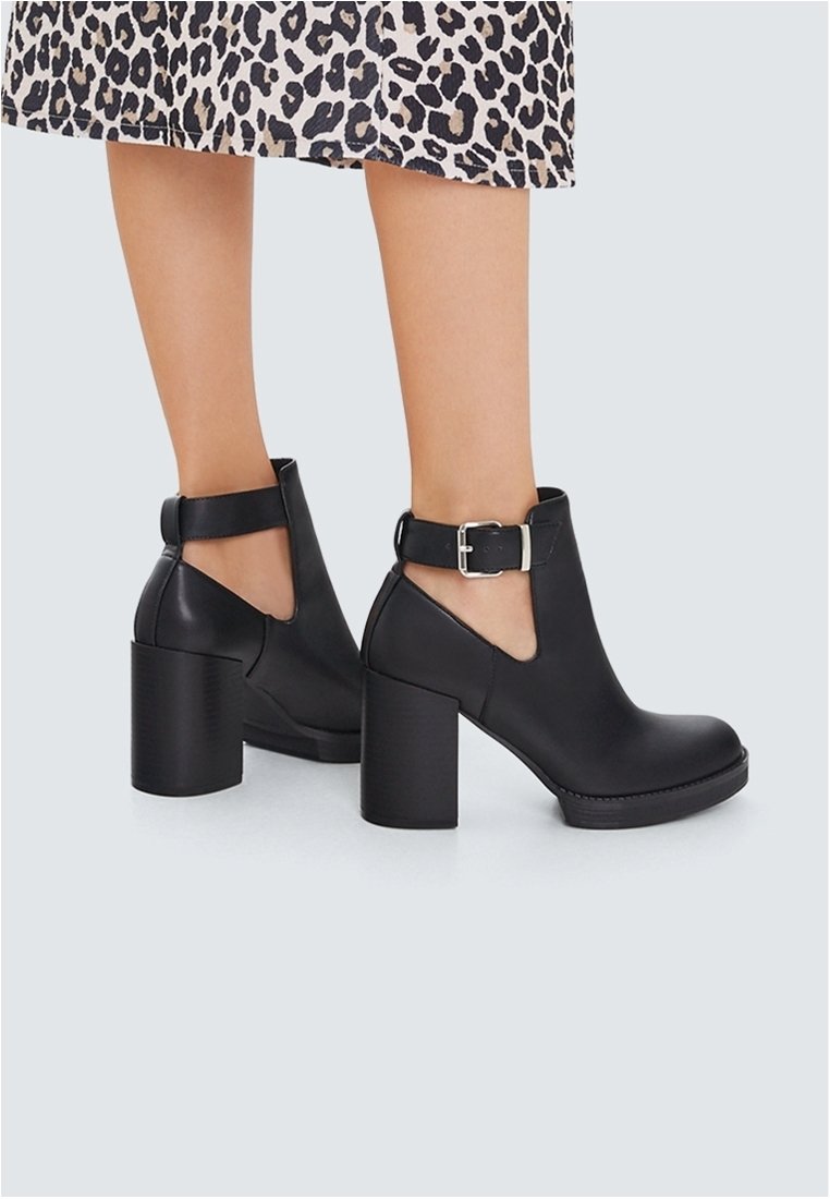 Bottines en cuir noir à talons blocs et découpes latérales, attachées par une bride à boucle argentée, portées avec une jupe imprimée léopard.