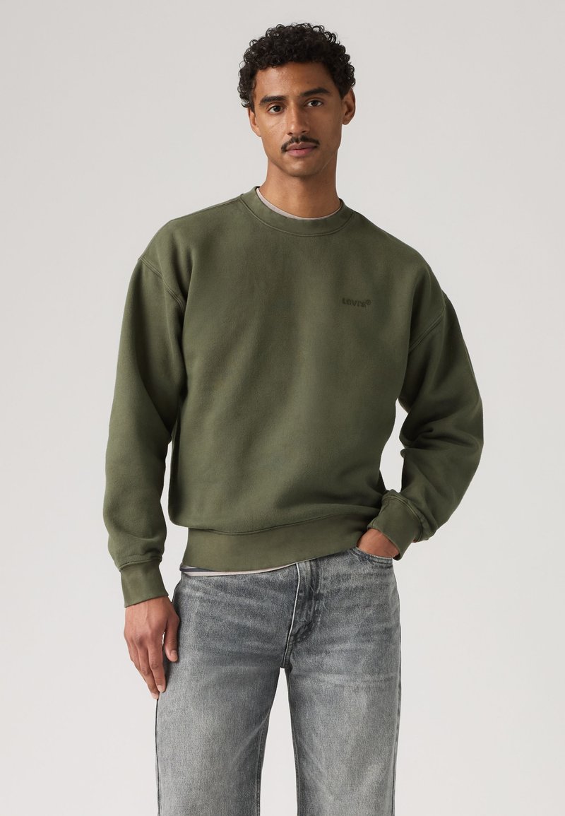 Uomo con capelli ricci e baffi indossa una felpa verde girocollo e jeans blu sbiaditi, in piedi con una mano in tasca su sfondo semplice.