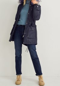 Doudoune marine avec capuche en fausse fourrure, taille ajustable par cordon, fermeture éclair avant et poches à boutons-pression ; associée à un pull bleu, un jean foncé et des baskets beige.