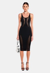 Schwarzes, enges Kleid mit transparenten Einsätzen, dünnen Trägern und knielanger Länge. Glatte Textur, ergänzt durch schwarze Riemchen-Heels und eine silberne Tasche.
