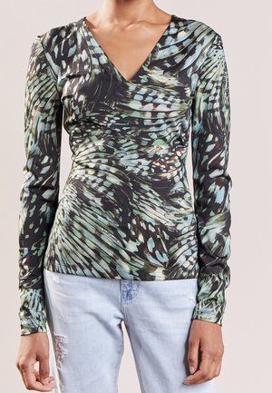 Kobieta nosząca dopasowaną bluzkę z długim rękawem w zielono-czarny wzór piór oraz jasnoniebieskie podarte dżinsy, stojąca na jednolitym tle.