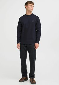 Marineblå genser med rund hals, strukturert stoff, med en liten logo på brystet. Kombinert med svarte bukser og brune mønstrede sko.