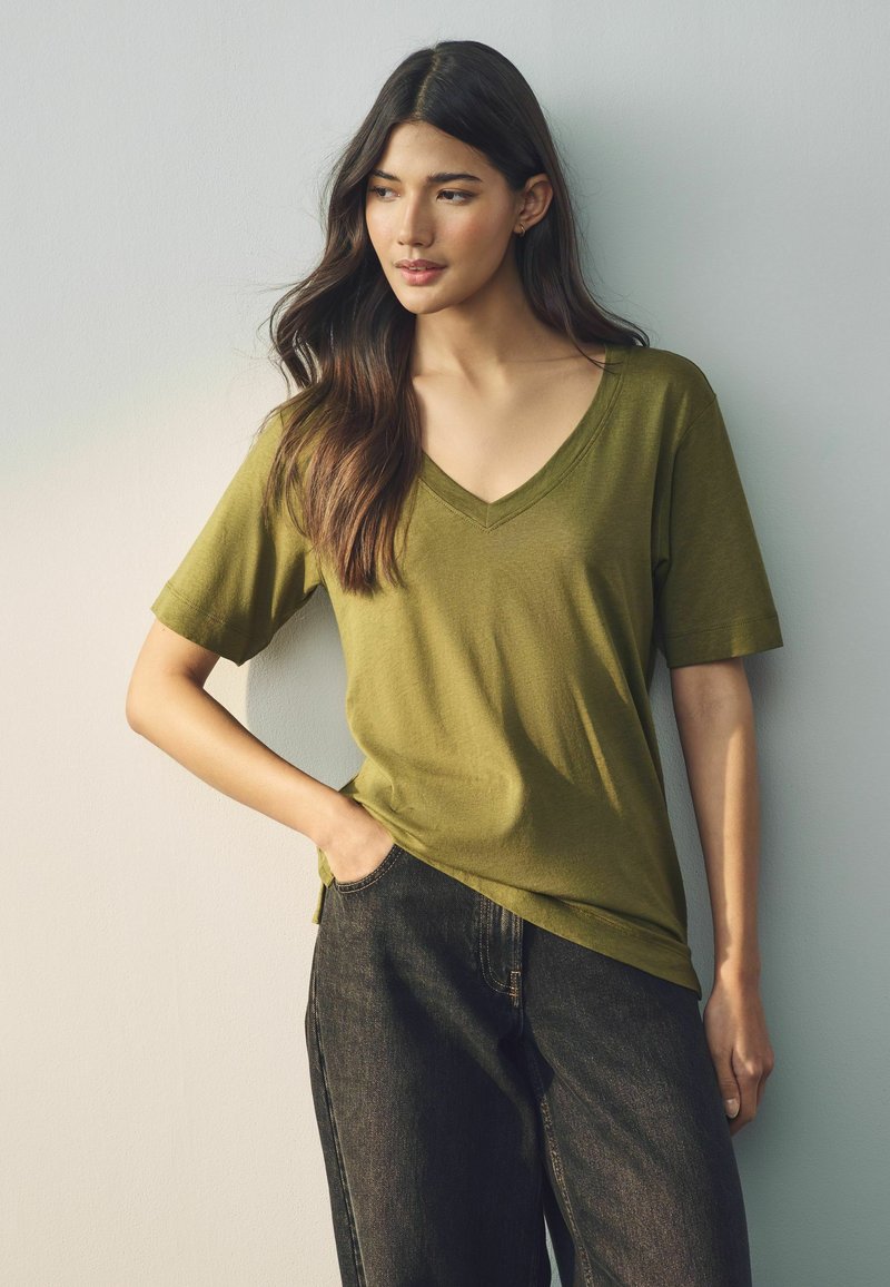Olijfgroene T-shirt met korte mouwen en V-hals van zachte stof, gecombineerd met zwarte denim jeans. Het T-shirt heeft een relaxte pasvorm en zijsplitten.