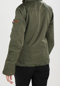Veste isolante vert olive avec une poche zippée sur le bras gauche, des accents à pression, une texture douce et un col montant.