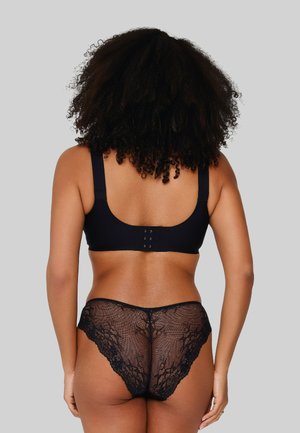 Donna con capelli ricci vista di spalle che indossa un reggiseno nero e biancheria intima in pizzo nero su uno sfondo grigio uniforme.