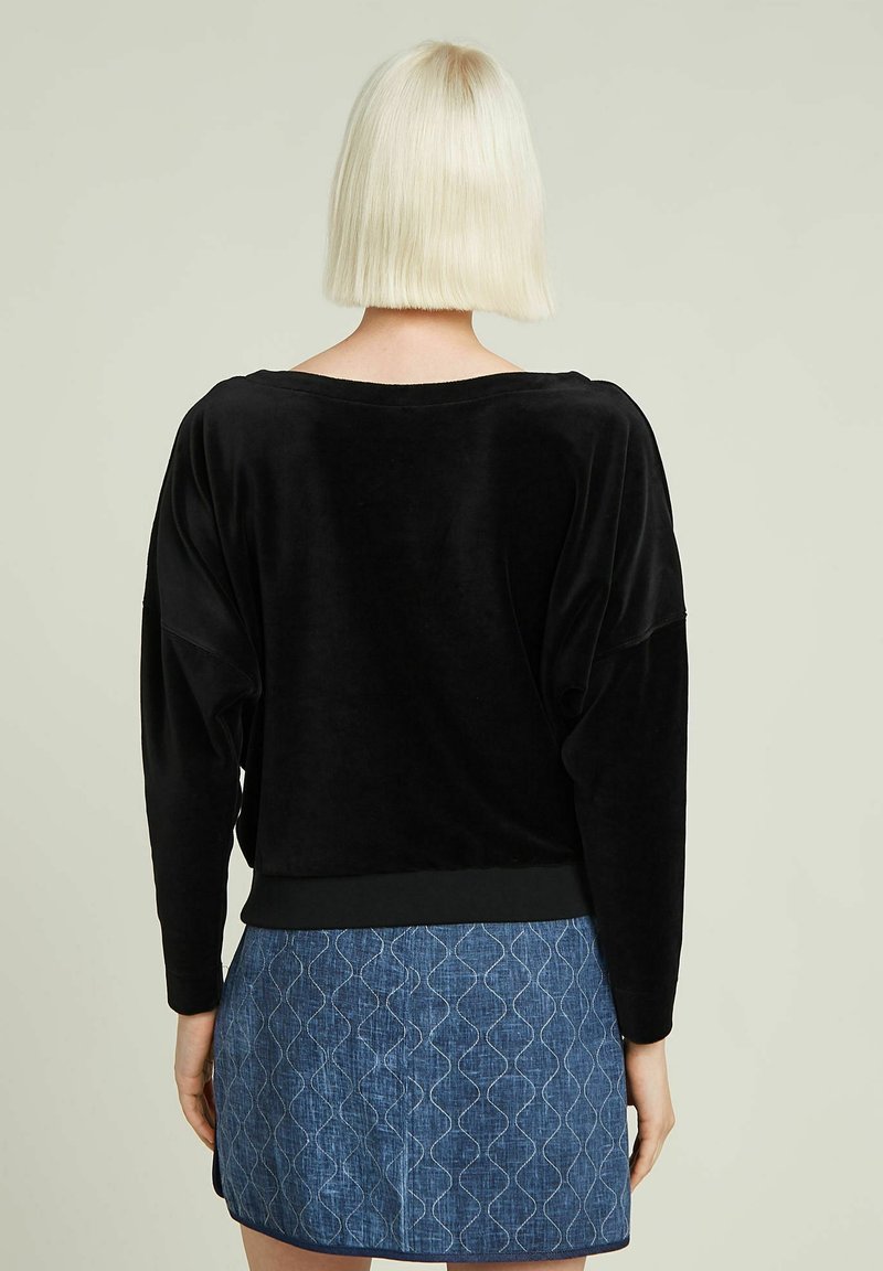 Schwarzer Samt-Pullover mit oversize Passform, überschnittenen Schultern und geripptem Saum; kombiniert mit einem blauen Denim-Rock mit wellenförmigem Muster.