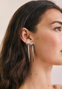 Silberne Creolen mit elongated Anhängern, die ein geknotetes Design und eine strukturierte Oberfläche aufweisen und am Ohrläppchen hängen.